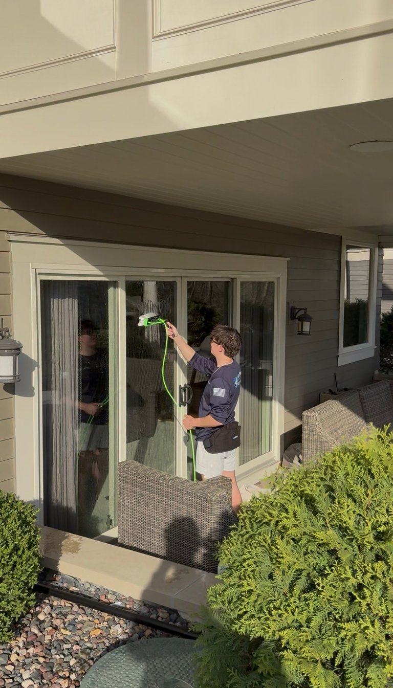 Person watering plants with green hose on patio near glass doors and landscaped bushes